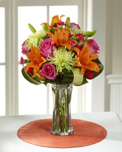 Hot pink bi-colored roses, orange Asiatic Lilies, green Fuji Chrysanthemums, hot pink mini carnations, tropical leaves, and lush greens are brought together to impress, presented in a clear glass vase. Bright, beautiful, and ready to get the party started, this bouquet creates a unique surprise to absolutely delight your recipient with each sunlit bloom.
<ul>
<li>Our florists hand-design each arrangement, so colors, varieties and containers may vary due to local availability</li>
</ul>